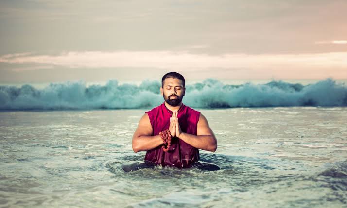 Acharya Dr. Ishan Shivanand Ji standing in the ocean with hands folded in prayer, waves rising behind him